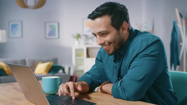 Portrait of Handsome Young Man Using Laptop Computer at Home, Watching and Laughing at Content. Happy Hispanic Man Works on Computer in a Cozy Living Room.