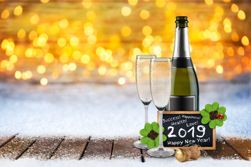 champagne bottle and glasses four leaf clover ladybug cork and blackboard happy new year 2019 greetings in front of bright golden warm bokeh lights snowy snow background wooden floor