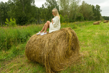Naklejka na ściany i meble A pretty blonde woman in a In a white knitted woolen jacket sits on a large round stack of dry hay collected on a summer or autumn day against a background of green grass at half a turn position