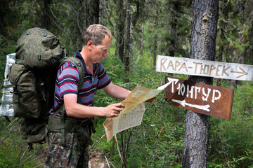 Tourist with a map near the signs. Inscriptions Tungur village, Kara Turek mountain. A man traveler got lost in a hike and is looking for a way on the map. - obrazy, fototapety, plakaty