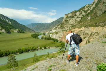 Young man photographer travels through the Altai with the camera on the tripod takes a shot of the snow-covered mountains and the rocks with turquoise winding river Katun. Photo tour.
