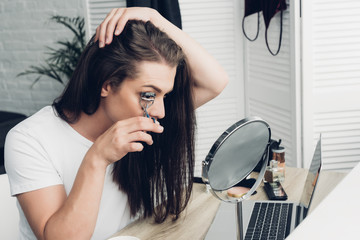 young transgender man doing makeup with lash curler at home