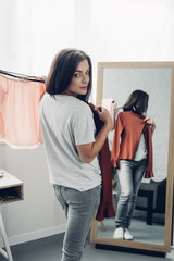 young transgender man trying on female shirts in front of mirror and looking at camera