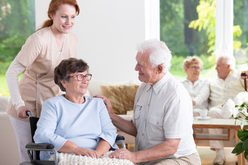Smiling elderly man visiting senior woman in the wheelchair while nurse supporting her