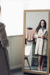 young transgender man in trench coat looking at mirror at home