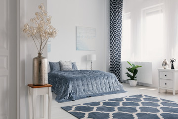 Flowers in gold vase in white bedroom interior with patterned carpet in front of blue bed. Real photo