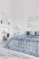 Plants and lamp next to blue bed in white minimal bedroom interior with posters. Real photo