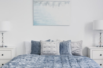 Poster above blue bed with pillows in white simple bedroom interior with lamps on cabinets. Real...