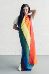 attractive young transgender man covering with pride flag and looking at camera in front of white...