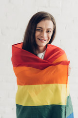 happy transgender man covering with pride flag and looking at camera in front of white brick wall