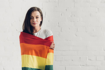 young transgender man covering with pride flag and looking at camera in front of white brick wall