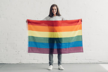 young transgender woman holding pride flag in front of white brick wall