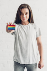 young transgender man holding card with pride flag in front of white brick wall