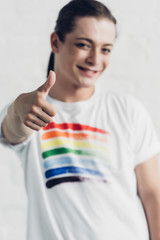 young transgender woman in white t-shirt with pride flag showing thumb up in front of white brick...