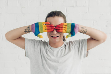 smiling transgender man covering eyes with hands painted in colors of pride flag in front of white...