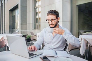 Obraz premium Concentrated man sits at table and works on laptop. He hold cup of tea on hand. There are opened notebook and pen at table. Businessman works.
