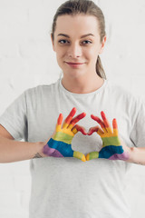 smiling young transgender man making heart sign with hands in colors of pride flag in front of...