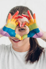 young transgender man looking at camera while making heart sign with hands in colors of pride flag...