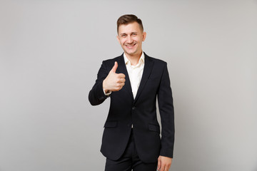 Portrait of successful young business man in classic black suit white shirt showing thumb up isolated on grey wall background in studio. Achievement career wealth business concept. Mock up copy space.