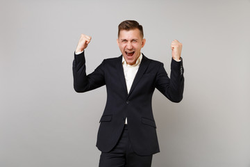 Portrait of overjoyed young business man in classic black suit, shirt screaming doing winner gesture isolated on grey wall background. Achievement career wealth business concept. Mock up copy space.