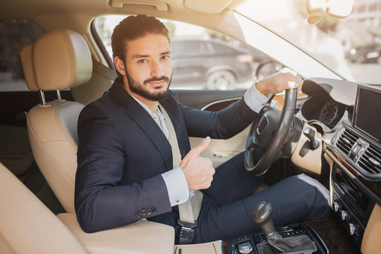 Nice picture of young businessman sitting in luxury car and holding big thumb up. He hold one hand on steering wheel. Young man recommends this car.