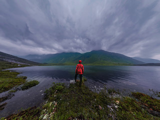 Fototapeta na wymiar man stands by lake in red jacket in cloudy weather panorama