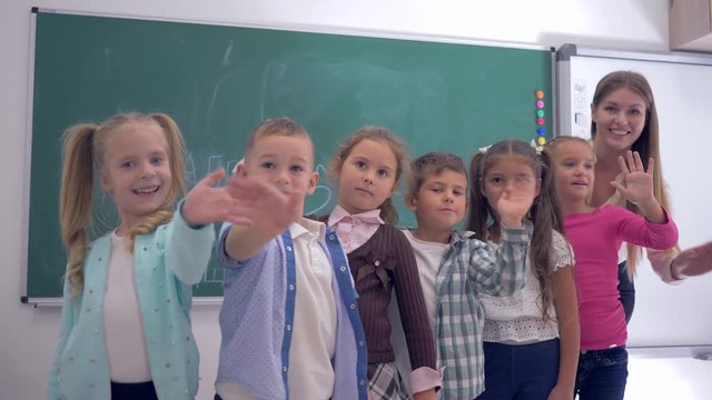 group of elementary school children with a young teacher waving hands on background of blackboard in classroom
