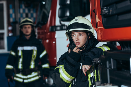 Selective Focus Of Female Firefighter Talking On Portable Radio Set With Colleague Behind At Fire Department