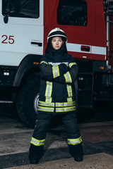 Naklejka premium female firefighter in helmet with arms crossed standing at fire station with truck behind
