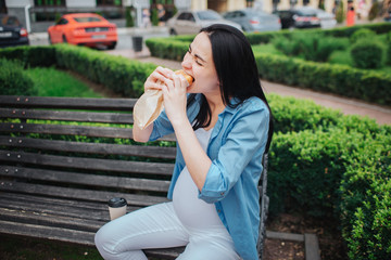 Portrait of a happy black hair and proud pregnant woman in a city in the background. Hungry expectant mother is listening to music in the park with an unborn child and eating a pie or sandwich. Hungry