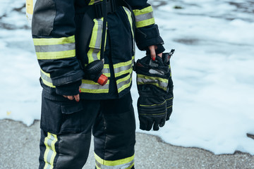 partial view of female firefighter in fireproof uniform with gloves in hand standing on steet with foam