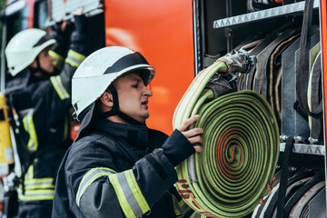 male firefighter in helmet putting water hose into truck on street