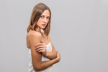 Naklejka premium Young freckled model stands and poses on camera. She keeps hands crossed. Young woman is serious and concentrated. She whas white towel across her body. Isolated on grey background.