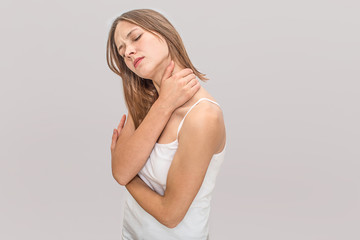 Naklejka premium Young woman feels pain in neck. She keeps eyes closed and hold hands together. Model wears white t-shirt. Isolated on grey background.