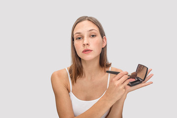 Young freckled model poses on camera. She holds pallete of eye shadows and use brush for that. Model looks on camera. She is cool. Isolated on grey background.