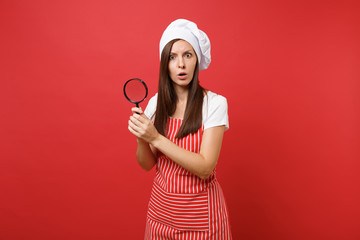 Housewife female chef cook or baker in striped apron white t-shirt toque chefs hat isolated on red wall background. Housekeeper woman hold and look through magnifying glass. Mock up copy space concept