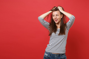 Portrait of attractive young woman in striped clothes standing, clinging to head, looking aside isolated on bright red wall background. People sincere emotions, lifestyle concept. Mock up copy space.