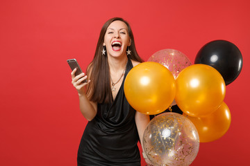 Overjoyed cheerful woman in little black dress hold air balloons using mobile phone while celebrating isolated on red background. Valentine's Day, Happy New Year birthday mockup holiday party concept.