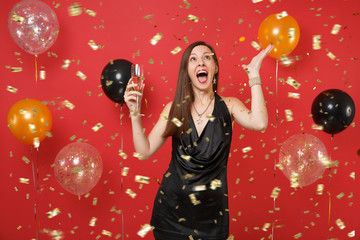 Excited young woman in little black dress celebrating, holding glass of champagne and catching confetti on bright red background air balloons. Happy New Year, birthday mockup holiday party concept.