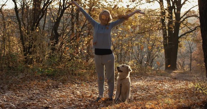 Young woman with her dog retriever tossing up leaves in autumn forest park