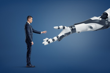 A tiny businessman stands in a side view and lifts his hand to touch a giant robotic arm.
