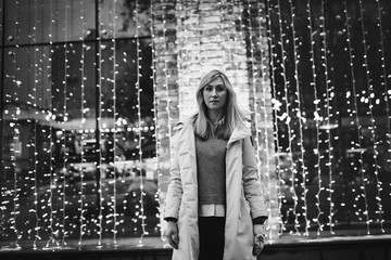Winter portrait of young woman walking in snowy city decorated for Christmas and New Year holidays, wearing in a white coat with fur hood. Christmas lights on background. Black and white photo.