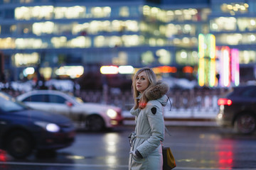 Beautiful young woman with big eyes and long blond hair in jacket with hood stands on evening city street.