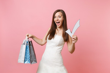Excited bride woman in wedding dress holding check mark, multi colored packages bags with purchases after shopping isolated on pink background. Wedding to do list. Organization of celebration concept.