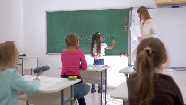 schoolgirl writes an simple example on the chalkboard near educator on math lesson, primary education