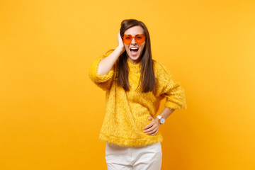 Excited happy young girl in fur sweater, white pants and heart orange glasses keeping hand near head isolated on bright yellow background. People sincere emotions, lifestyle concept. Advertising area.