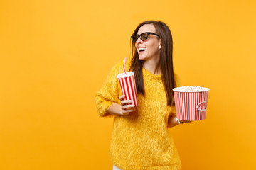 Joyful young woman in 3d imax glasses looking aside watching movie film hold bucket of popcorn, plastic cup of cola or soda isolated on yellow background. People sincere emotions in cinema, lifestyle.
