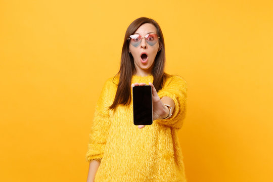 Shocked young woman in heart glasses showing on camera mobile phone with blank black empty screen isolated on bright yellow background. People sincere emotions, lifestyle concept. Advertising area.