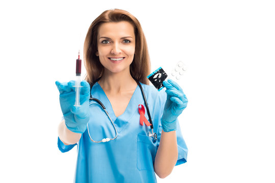 attractive smiling doctor with red ribbon holding syringe with blood and condom with pills isolated on white, world aids day concept
