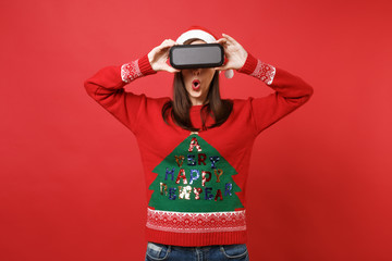 Amazed Santa girl with opened mouth covering eyes with portable wireless bluetooth music speaker isolated on red background. Happy New Year 2019 celebration holiday party concept. Mock up copy space.